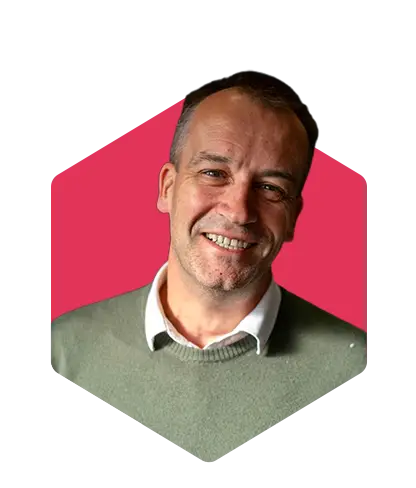Smiling man in a green sweater over a white collared shirt, portrait against a pink hexagonal background.